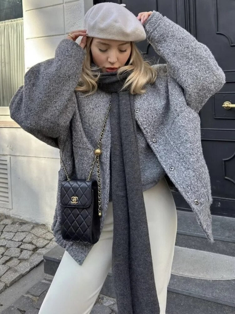 Frau in grauem Wollmantel, weißer Hose, schwarzer Tasche und beiger Baskenmütze vor Haustür.