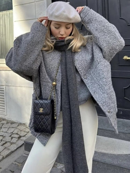 Frau in grauem Wollmantel, weißer Hose, schwarzer Tasche und beiger Baskenmütze vor Haustür.