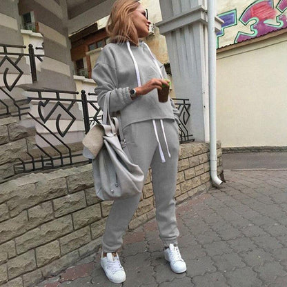Frau in grauem Jogginganzug, hält Getränk, trägt weiße Sneaker, lässiger Streetstyle-Look.