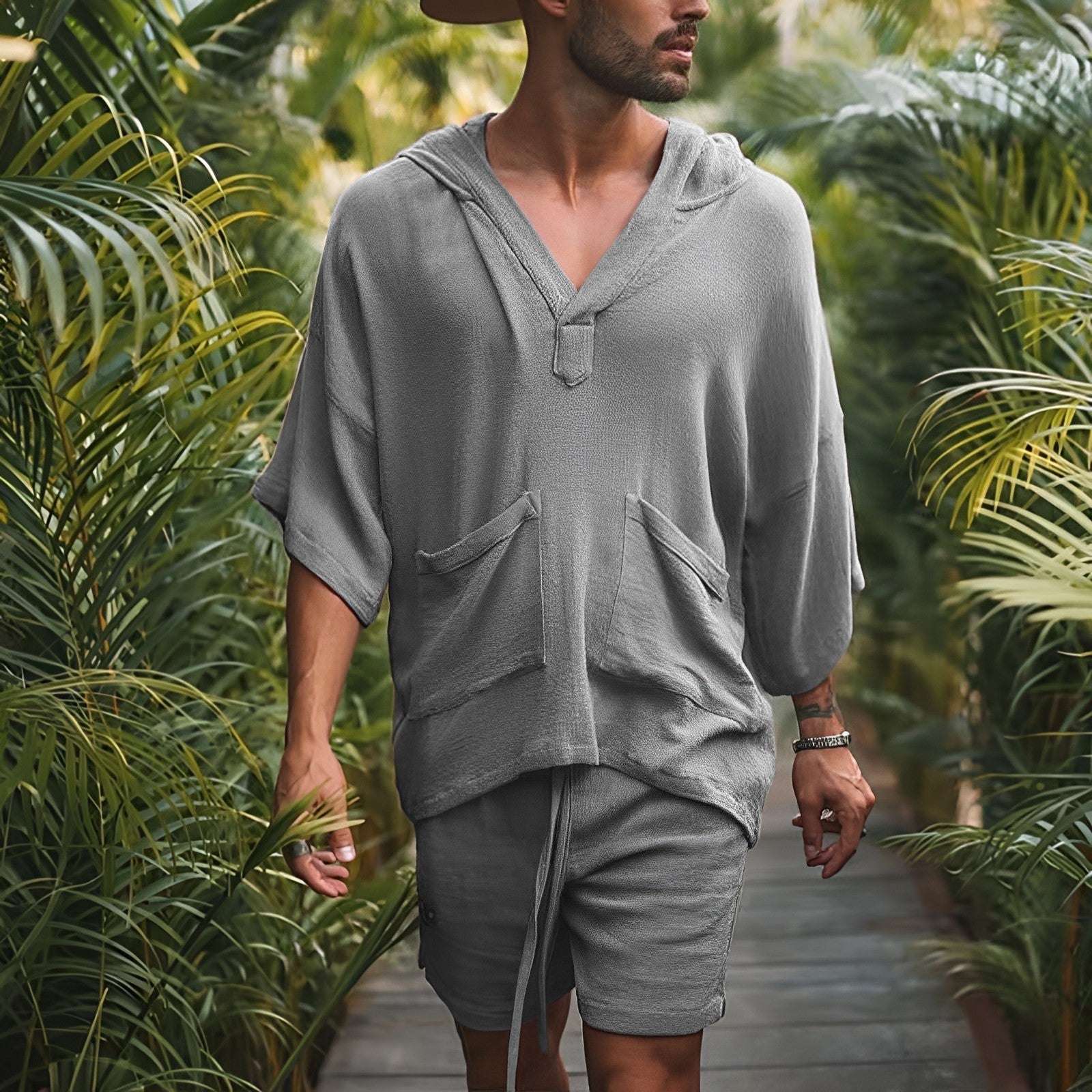 Mann in grauem Leinen-Hoodie und Shorts, lässige Sommermode, umgeben von tropischen Pflanzen.