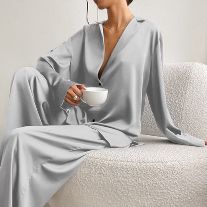 Frau in grauem Satin-Pyjama hält Tasse, sitzt auf weißem Sofa, elegantes Lounge-Outfit.