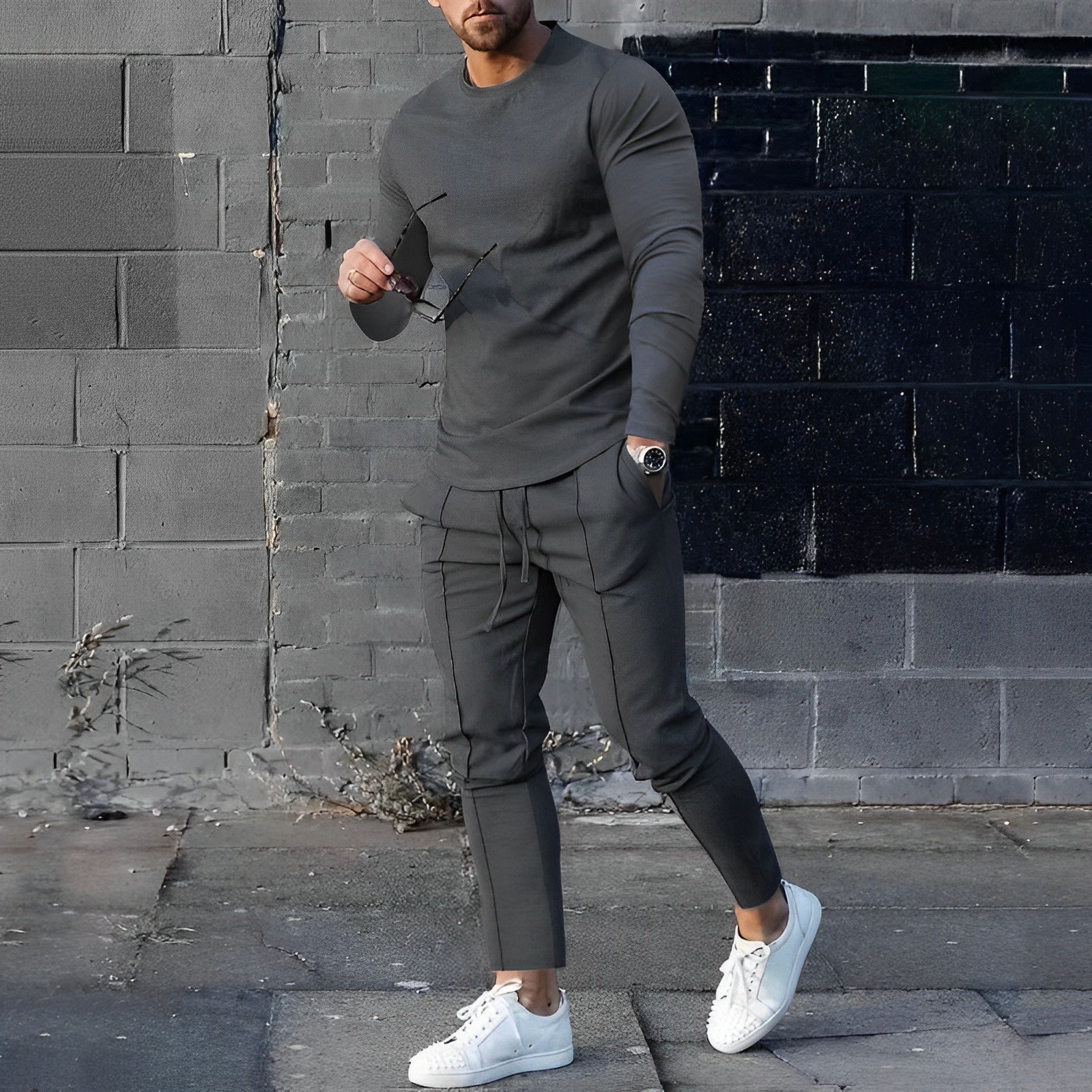 Mann in grauem Trainingsanzug und weißen Sneakers vor grauer Backsteinmauer.