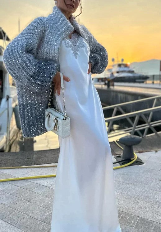 Elegantes weißes Abendkleid mit grauem Strickmantel und weißer Handtasche am Hafen bei Sonnenuntergang.