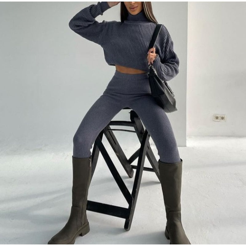 Frau in grauem Strickpullover und Leggings, schwarze Stiefel, sitzt auf Hocker, modisch, elegant.