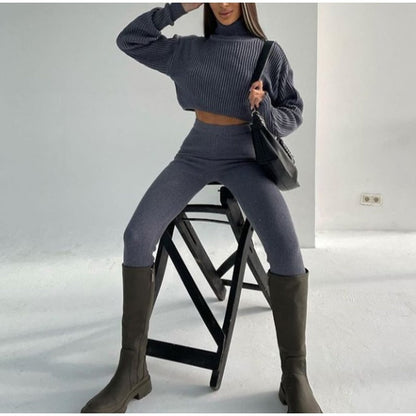 Frau in grauem Strickpullover und Leggings, schwarze Stiefel, sitzt auf Hocker, modisch, elegant.