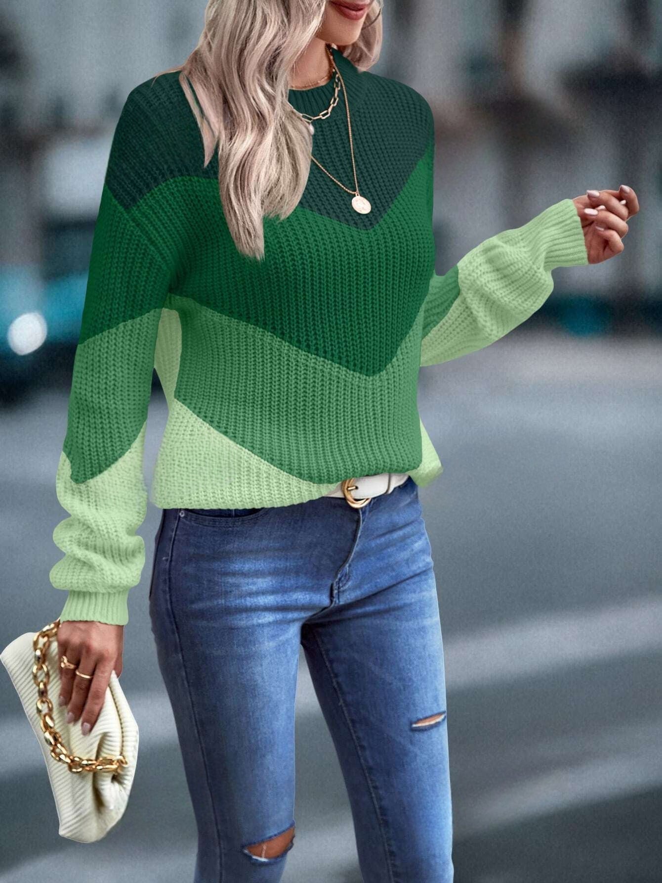 Frau in grünem Strickpullover, blaue Jeans, weiße Tasche, modisch, urbaner Hintergrund.