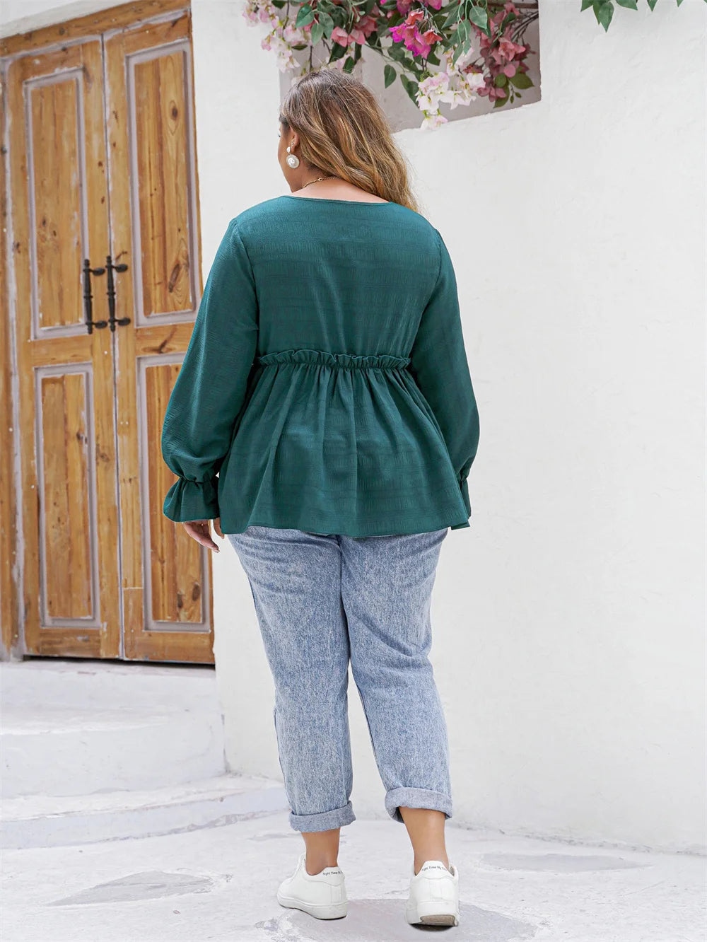 Frau in grüner Bluse und blauen Jeans, weiße Sneakers, vor Holztür und Blumen.