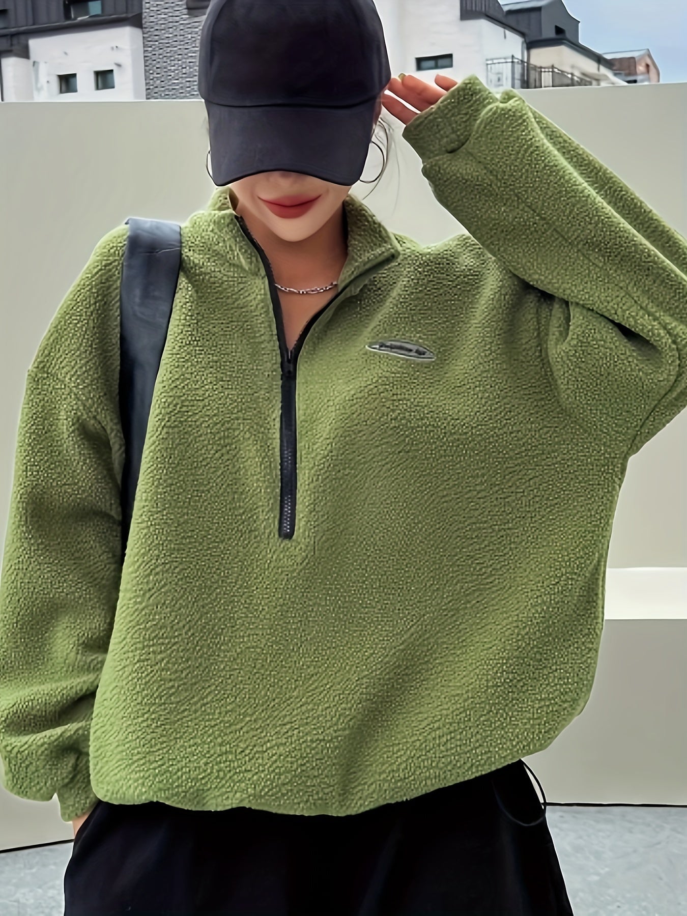 Frau in olivgrünem Fleece-Pullover mit Reißverschluss und schwarzer Kappe, urbaner Hintergrund.