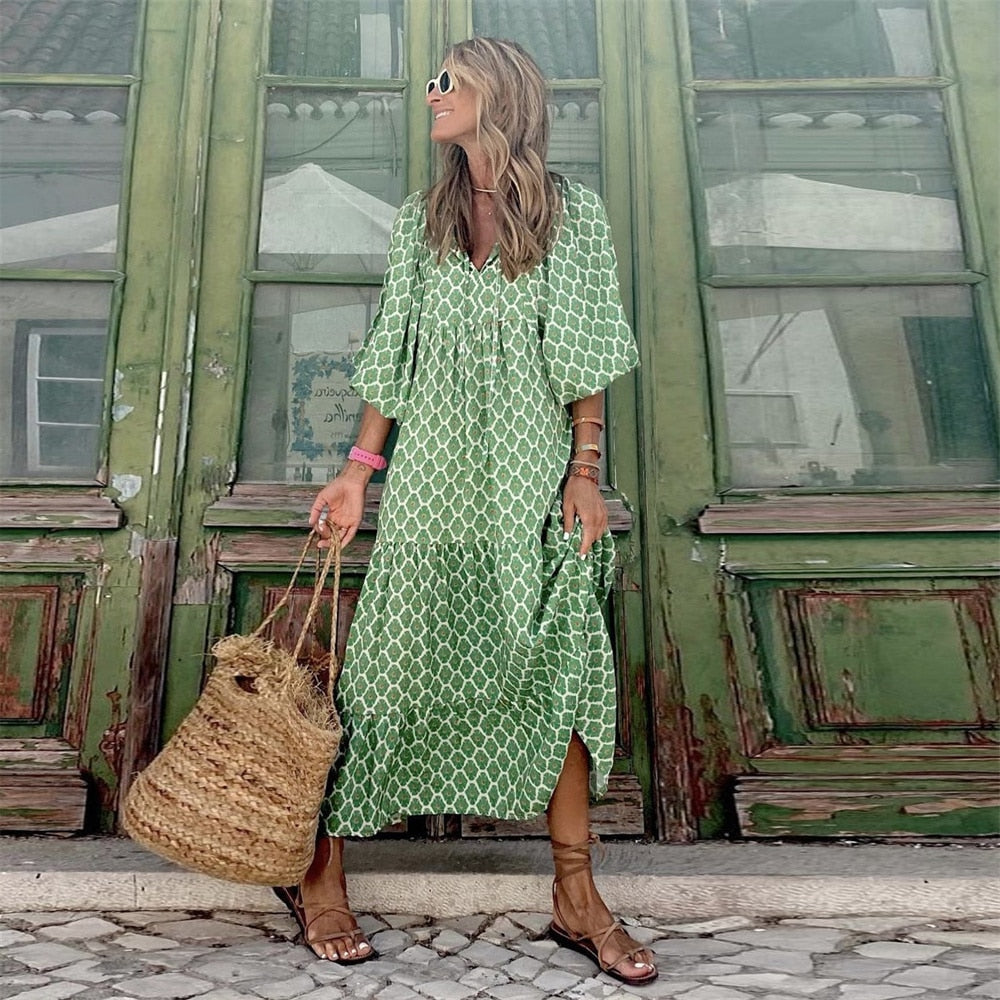 Frau in grünem Maxikleid mit geometrischem Muster, Strohtasche, Sandalen vor grüner Holztür.