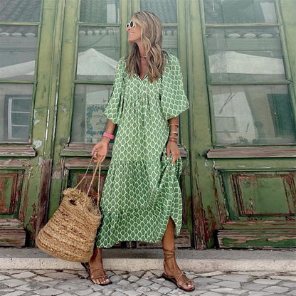 Frau in grünem Maxikleid mit geometrischem Muster, Strohtasche, Sandalen vor grüner Holztür.