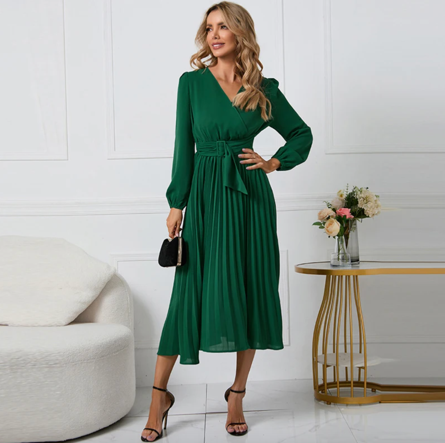 Frau in grünem Kleid mit langen Ärmeln, hält schwarze Clutch, steht neben Tisch mit Blumen.