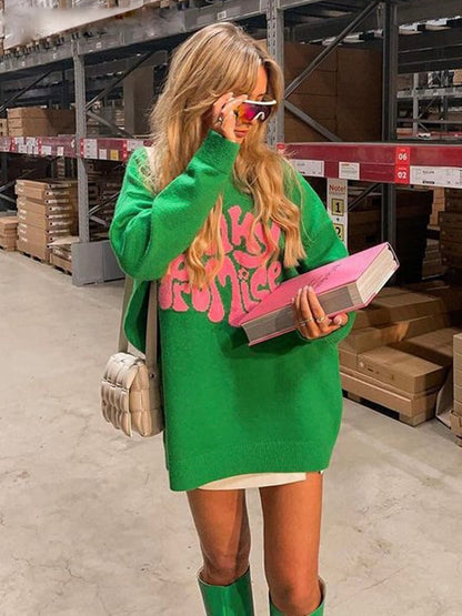 Frau in grünem Oversize-Pullover mit pinkem Schriftzug, hält Buch, trägt Sonnenbrille und Stiefel.