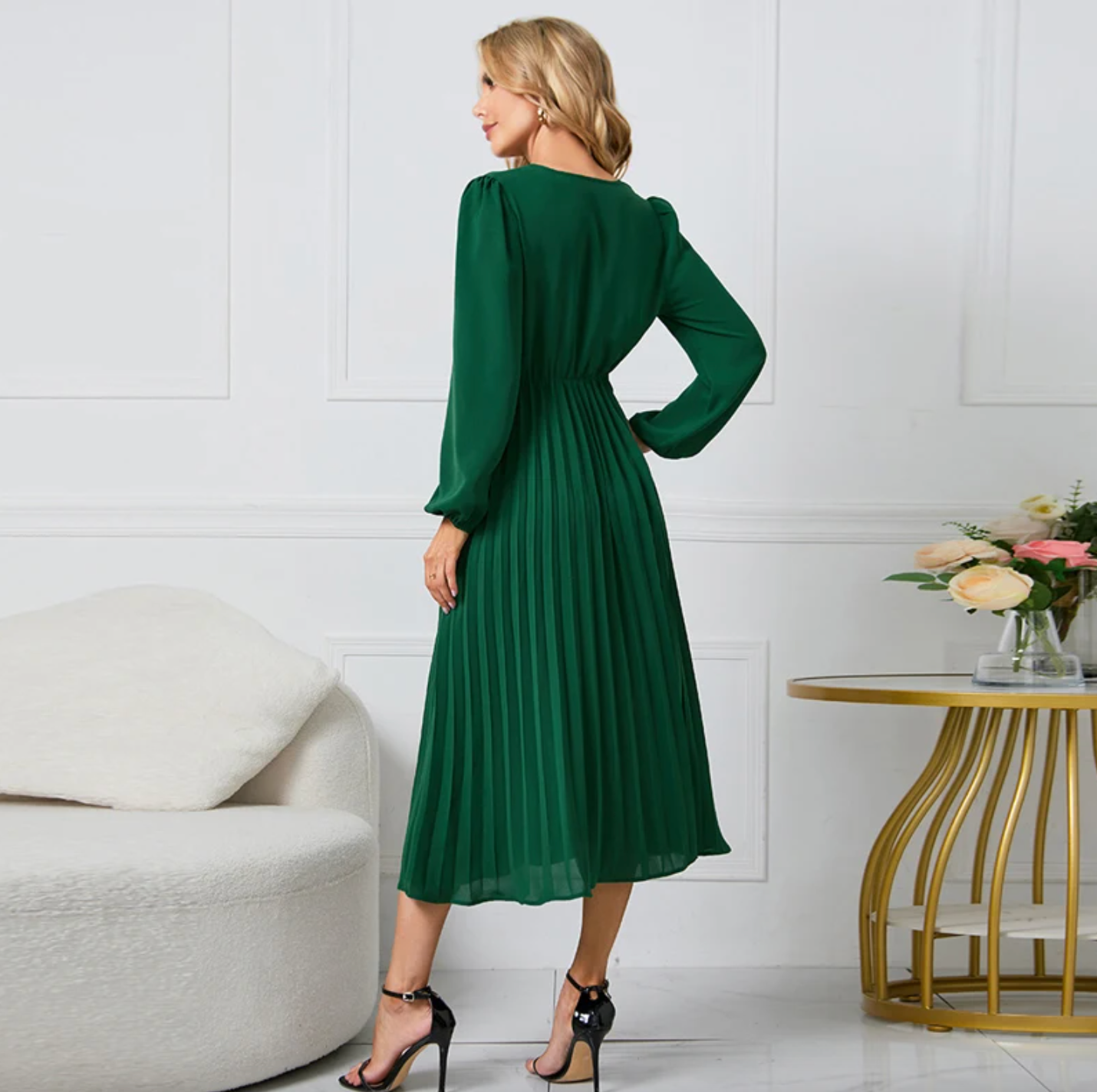 Frau in grünem, langem Kleid mit Falten, steht neben modernem Sofa und Tisch mit Blumen.