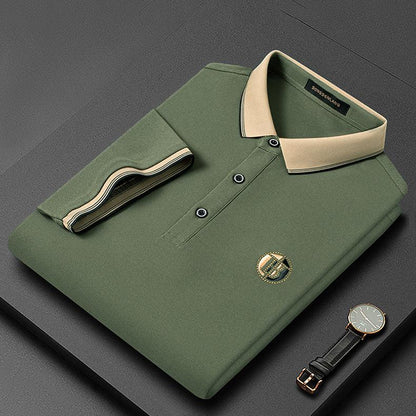 Grünes Herren-Poloshirt mit beigem Kragen, gefaltet, neben einer Armbanduhr auf grauem Hintergrund.