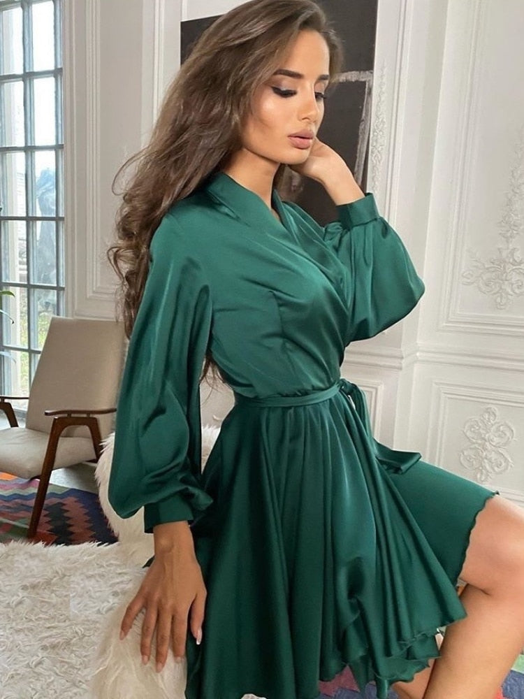 Frau in elegantem, grünem Satin-Kleid mit langen Ärmeln, sitzt stilvoll im Wohnzimmer.