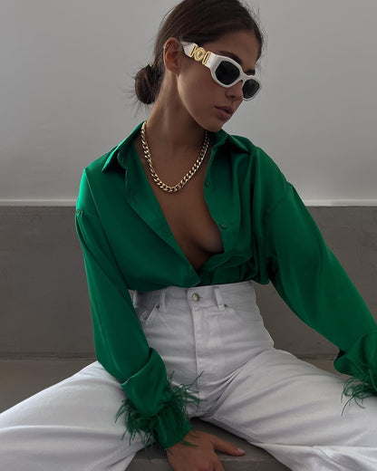 Frau in grüner Bluse und weißer Hose, trägt Sonnenbrille und goldene Halskette, modisch elegant.