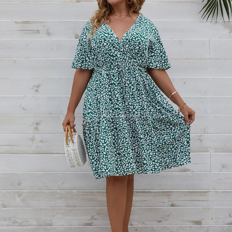 Frau in grünem Sommerkleid mit Blumenmuster, hält weiße Handtasche, vor Holzwand.