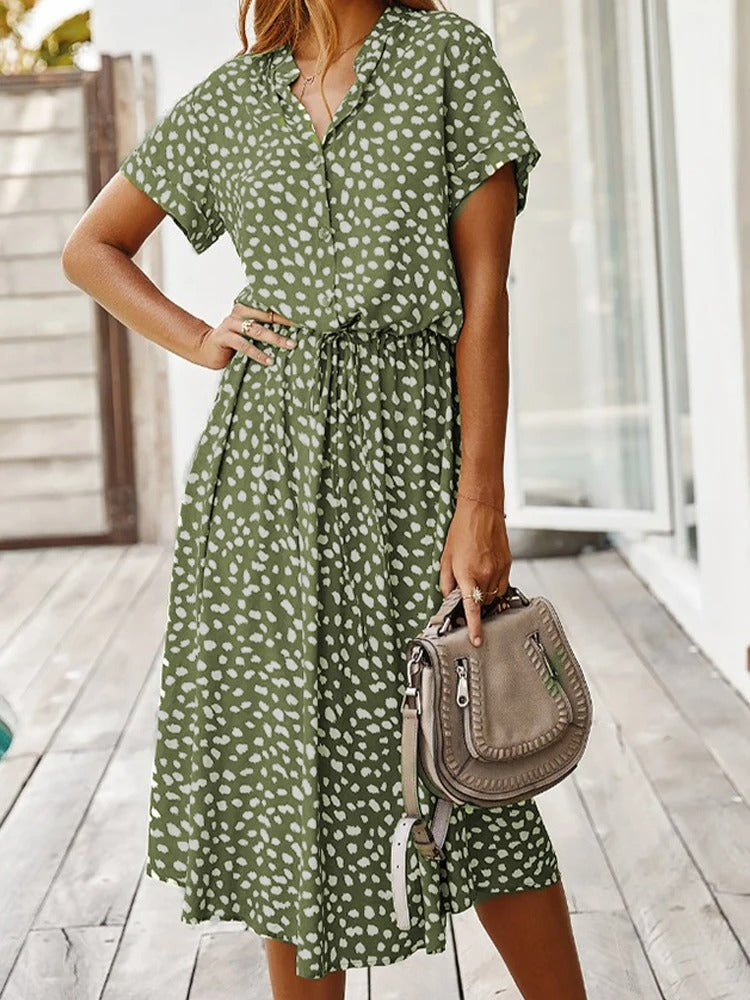 Frau in grünem Sommerkleid mit weißen Punkten, hält beige Handtasche, Holzterrasse im Hintergrund.