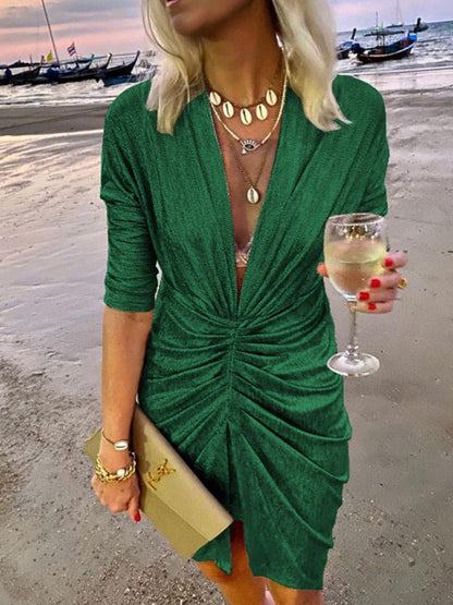 Frau in grünem Kleid am Strand, hält Weinglas, trägt goldene Accessoires, Abendmode.