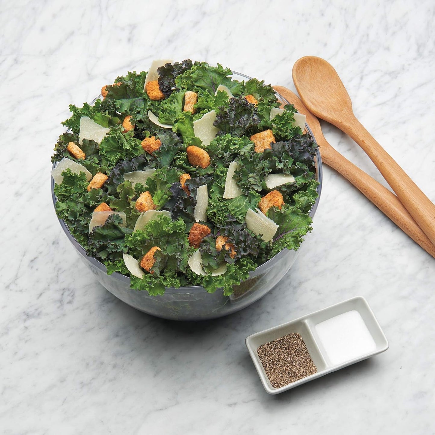 Grüner Salat mit Croutons und Parmesan in Glasschüssel auf Marmortisch, Holzlöffel daneben.