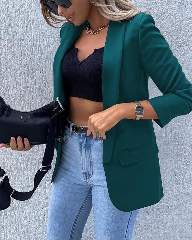 Frau in grünem Blazer, schwarzem Top, blauer Jeans, mit schwarzer Tasche und Sonnenbrille.