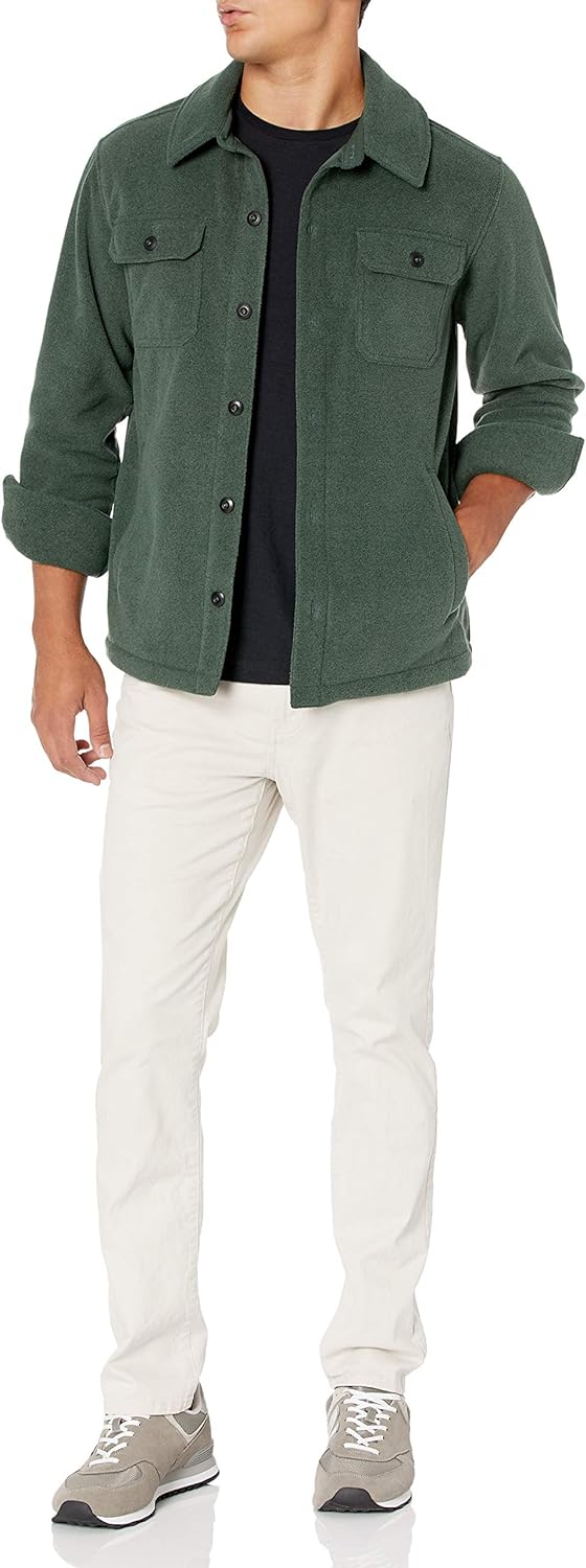 Mann in grüner Fleecejacke, schwarzem T-Shirt, weißen Hosen und grauen Sneakers stehend.