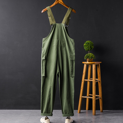 Grüne Latzhose aus Baumwolle, Unisex, mit Taschen, auf Holzkleiderbügel vor schwarzem Hintergrund.