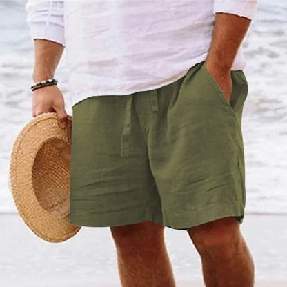 Mann in grüner Leinen-Shorts und weißem Hemd am Strand, hält Strohhut in der Hand.