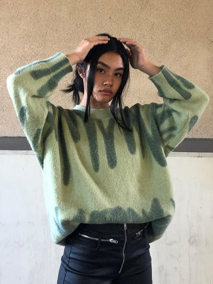 Frau in grünem Oversize-Pullover mit Muster, schwarze Hose, lässiger Look.
