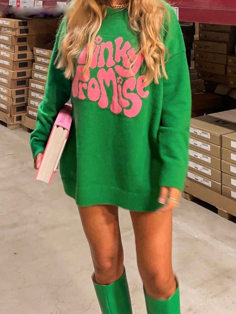 Frau in grünem Oversize-Pullover mit "Pinky Promise"-Aufdruck, hält ein Buch, trägt grüne Stiefel.