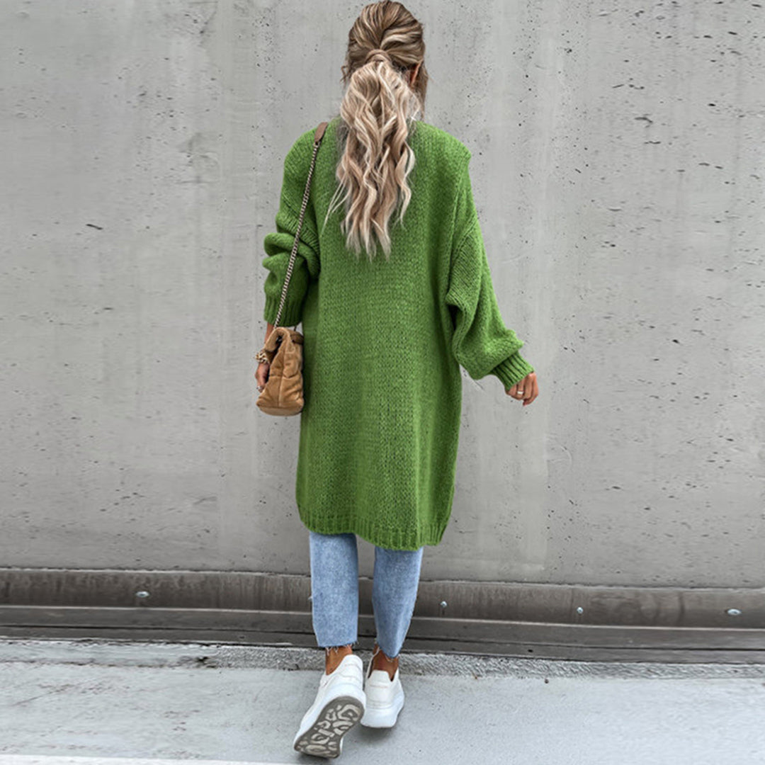 Frau in grünem Strickpullover, Jeans und weißen Sneakers vor Betonwand.