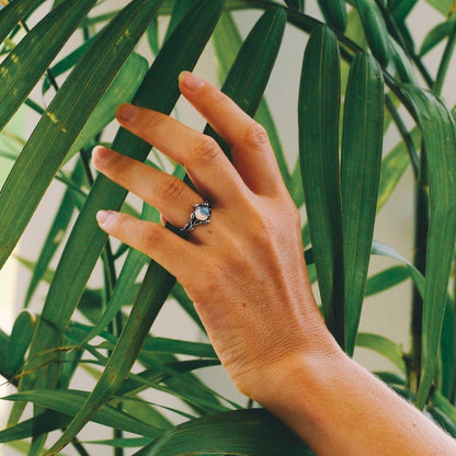 Hand mit Silberring und grünem Edelstein, umgeben von grünen Blättern, Schmuckfotografie.