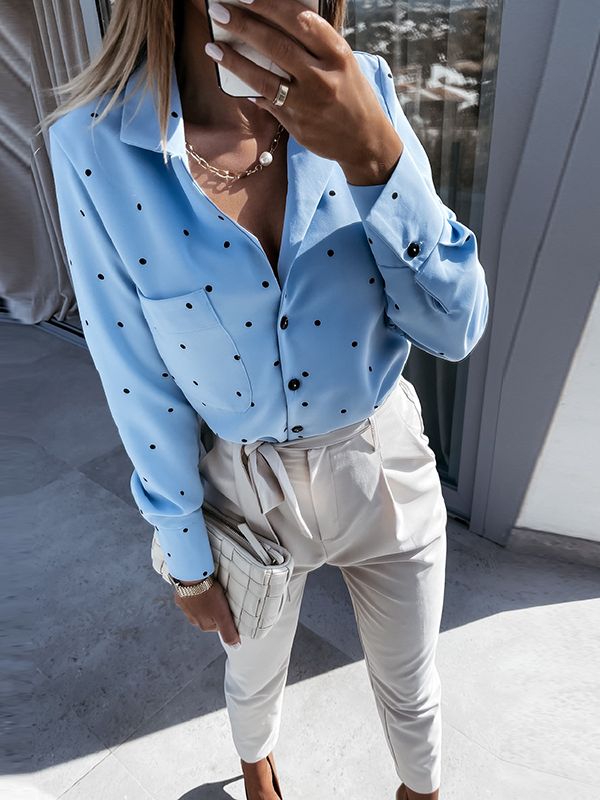 Frau in blauer gepunkteter Bluse und weißer Hose, hält weiße Clutch, modisch elegant.
