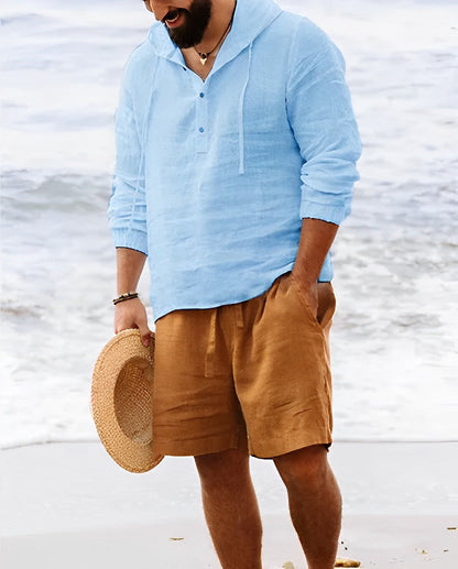 Mann am Strand in hellblauem Leinenhemd und braunen Shorts, hält Strohhut.
