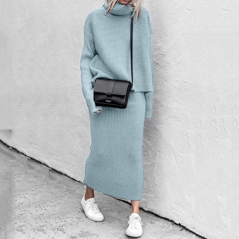 Frau in hellblauem Strickkleid mit schwarzer Umhängetasche und weißen Sneakers vor grauer Wand.