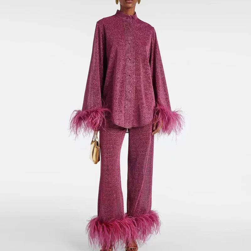 Modell i vinröd glittrig skjorta med ståkrage och byxor, båda med rosa fjäderkant, håller en liten guldfärgad väska. 1