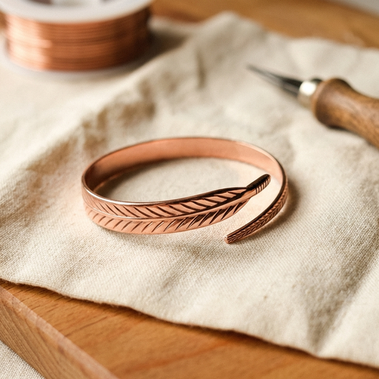 Koppararmband med graverat fjädermönster som ligger öppet på ett linnetyg, med en trådrulle och ett gravyrverktyg i bakgrunden. 1