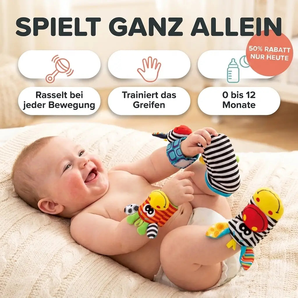 Leende bebis ligger p\u00e5 en stickad filt med rassel runt handlederna och randiga rasself\u00f6tter p\u00e5 f\u00f6tterna och greppar om foten. 1