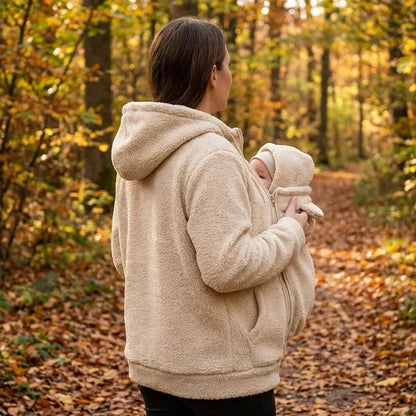 Kvinna i beige fleecehoodie sedd bakifrån och från sidan, bär en bebis i panelen på en skogsstig. 6