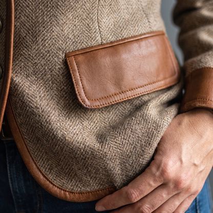 Närbild på en ficka med läderlock, läderkantad nederkant och ärmuppslag i konstläder mot sillbensmönstrad tweed. 5