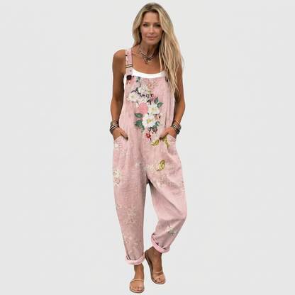 Hel figur framifrån: modell i hängselbyxorna över vitt linne, med blommönster, knäppta axelband, händerna i fickorna och sandaler. 2
