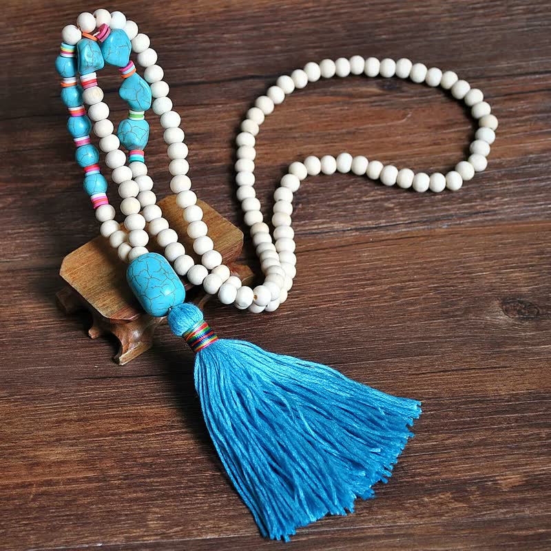 Mala-Kette mit blauen Quasten, weißen Perlen und türkisfarbenen Steinen auf Holzuntergrund.