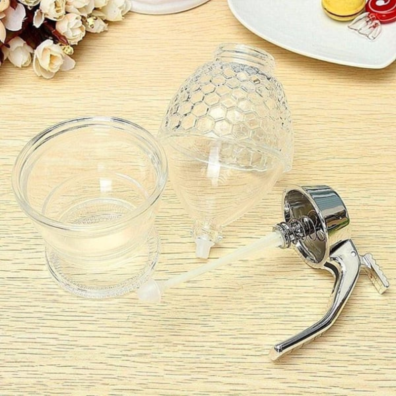 Honigspender aus Glas mit Metallgriff, transparent, bienenwabenförmig, auf Holztisch.