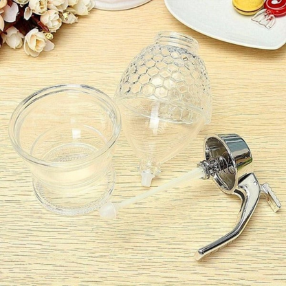 Honigspender aus Glas mit Metallgriff, transparent, bienenwabenförmig, auf Holztisch.