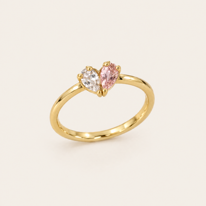 Guldfärgad ring sedd snett framifrån med en klar och en rosa droppformad sten som tillsammans bildar ett hjärta. 1