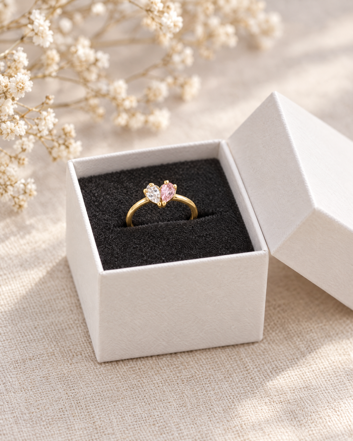 Guldfärgad ring med klar och rosa sten i en öppen vit ask med svart skuminlägg, intill torkade blommor. 3