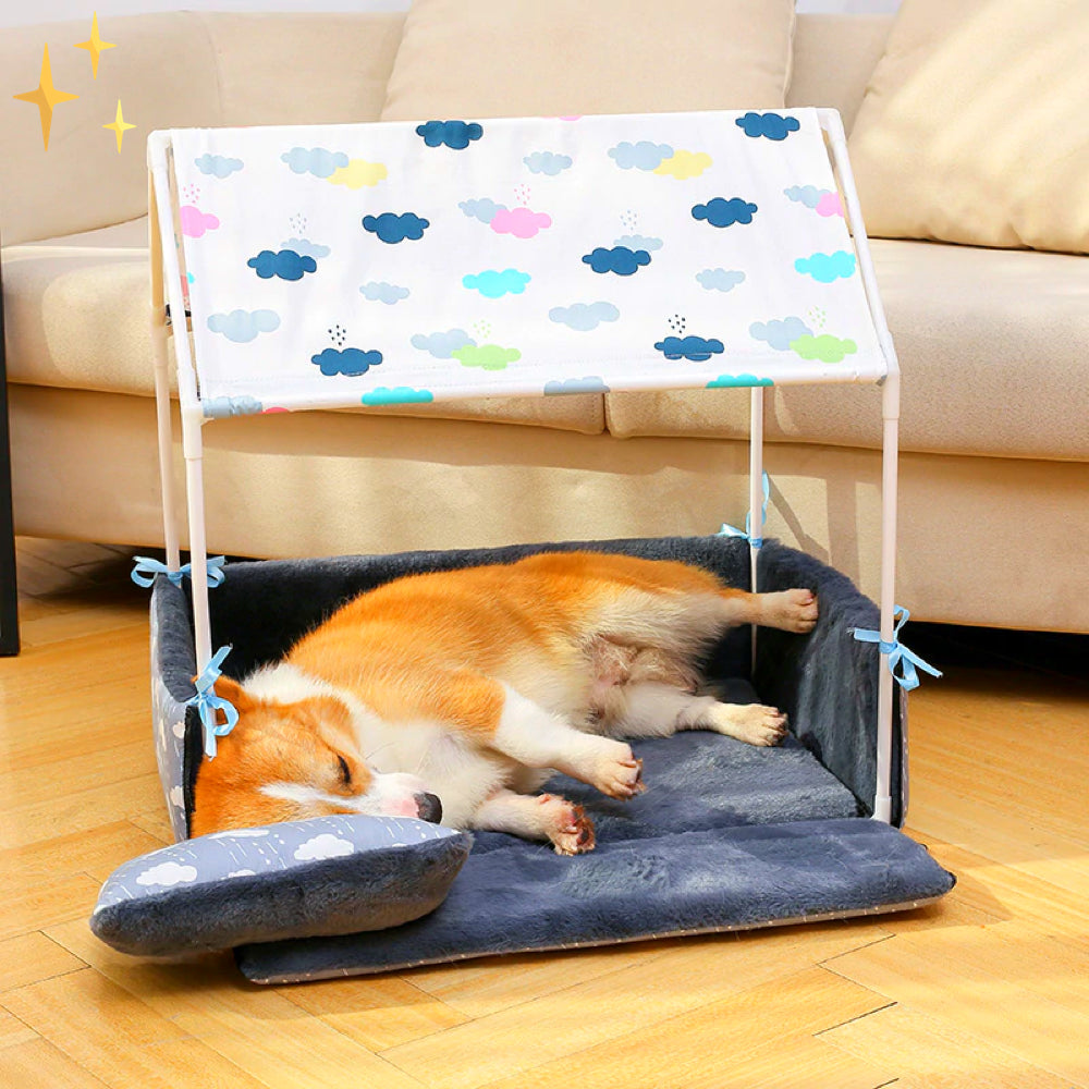 Schlafender Corgi in einem bunten Hundebett mit Dach auf Holzboden vor einem Sofa.