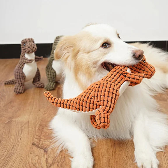 Hund spielt mit orangefarbenem Dinosaurier-Spielzeug aus Stoff auf Holzboden, zwei weitere Spielzeuge im Hintergrund.
