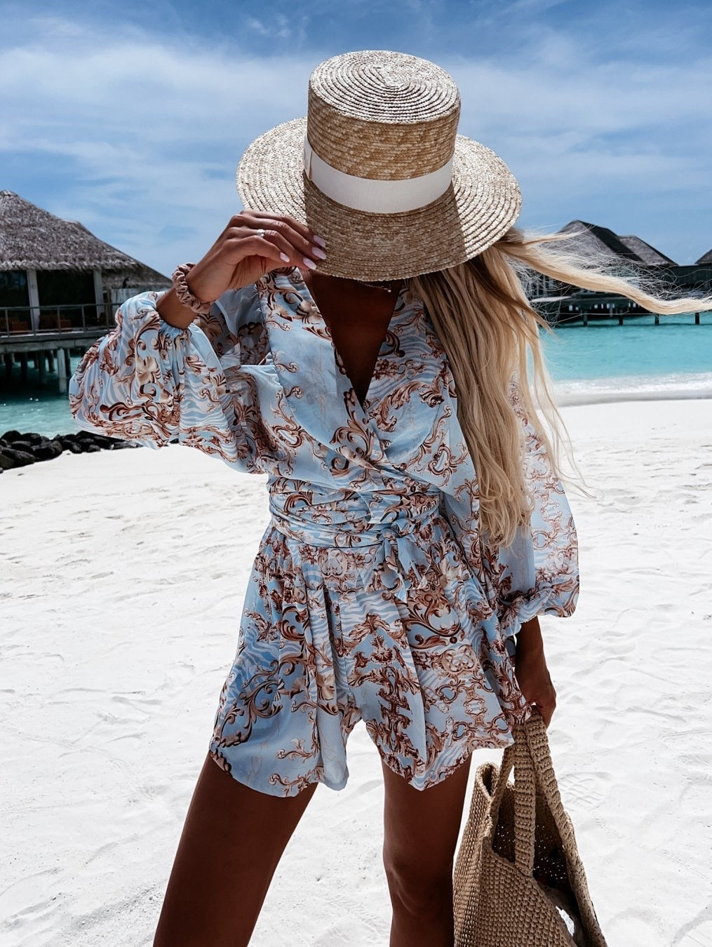 Frau in blauem gemustertem Sommerkleid und Strohhut am Strand mit Tasche, tropische Kulisse.