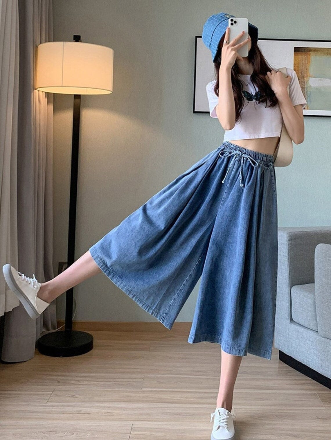 Frau in blauer Jeans-Culotte und weißem Crop-Top, trägt weißen Hut und Sneakers, moderne Mode.